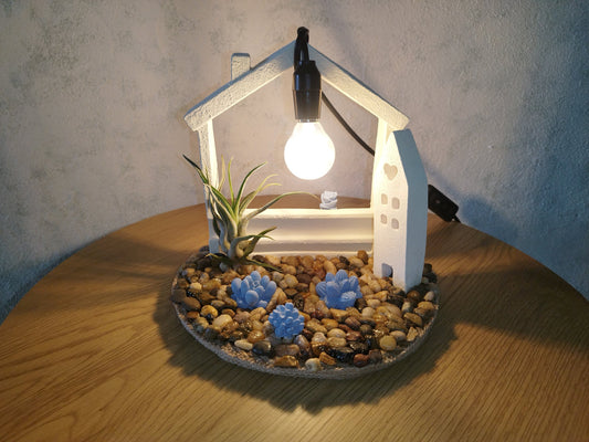 Decorative lamp shaped like a house with plants and rocks on a wooden table