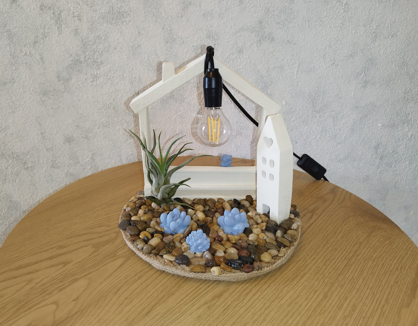 Decorative lamp with a house-shaped base, plants, and a light bulb on a wooden table