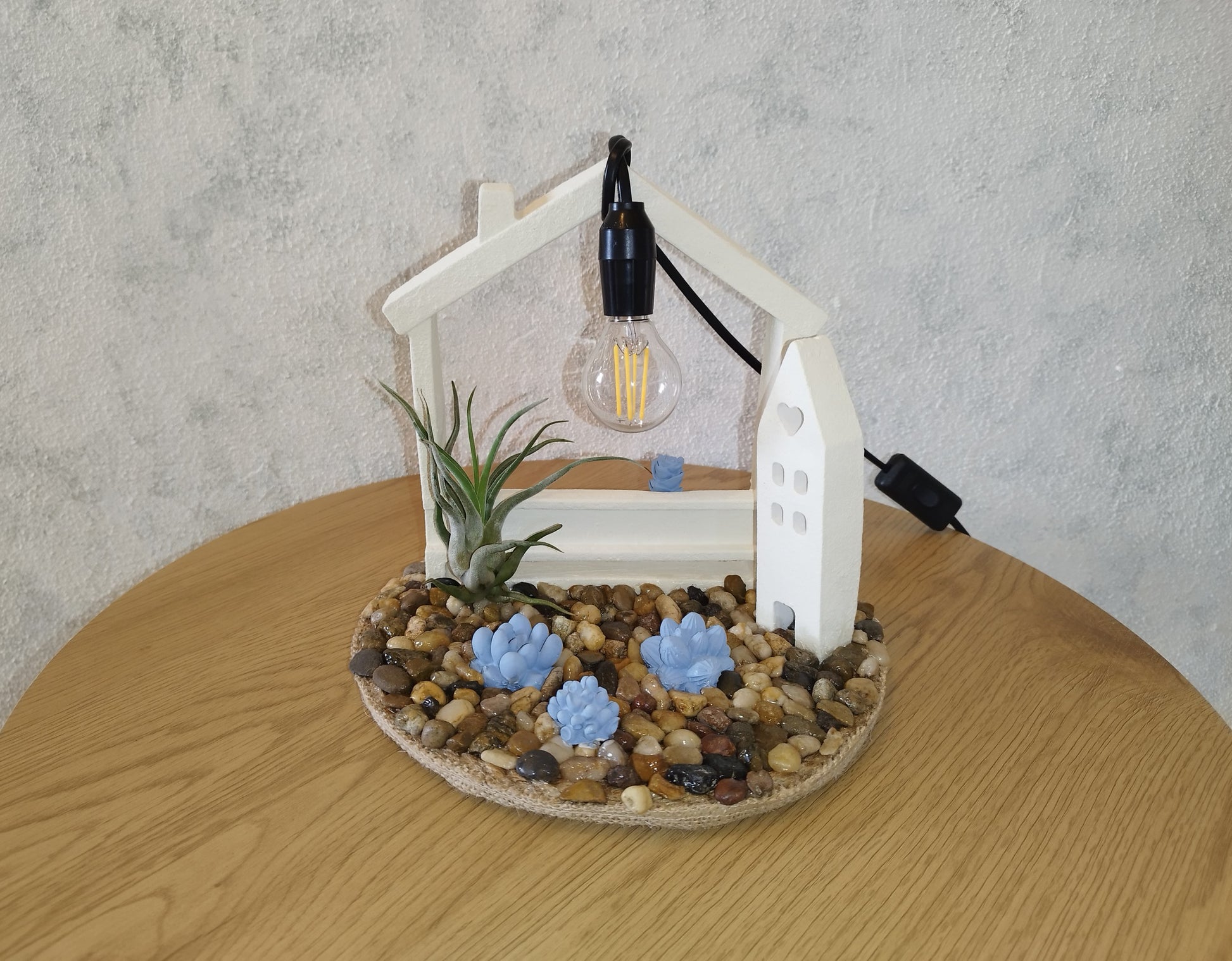Decorative lamp with a house-shaped base, plants, and a light bulb on a wooden table
