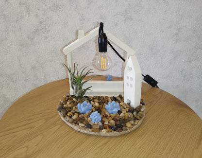 Decorative lamp with a house-shaped base, plants, and a light bulb on a wooden table